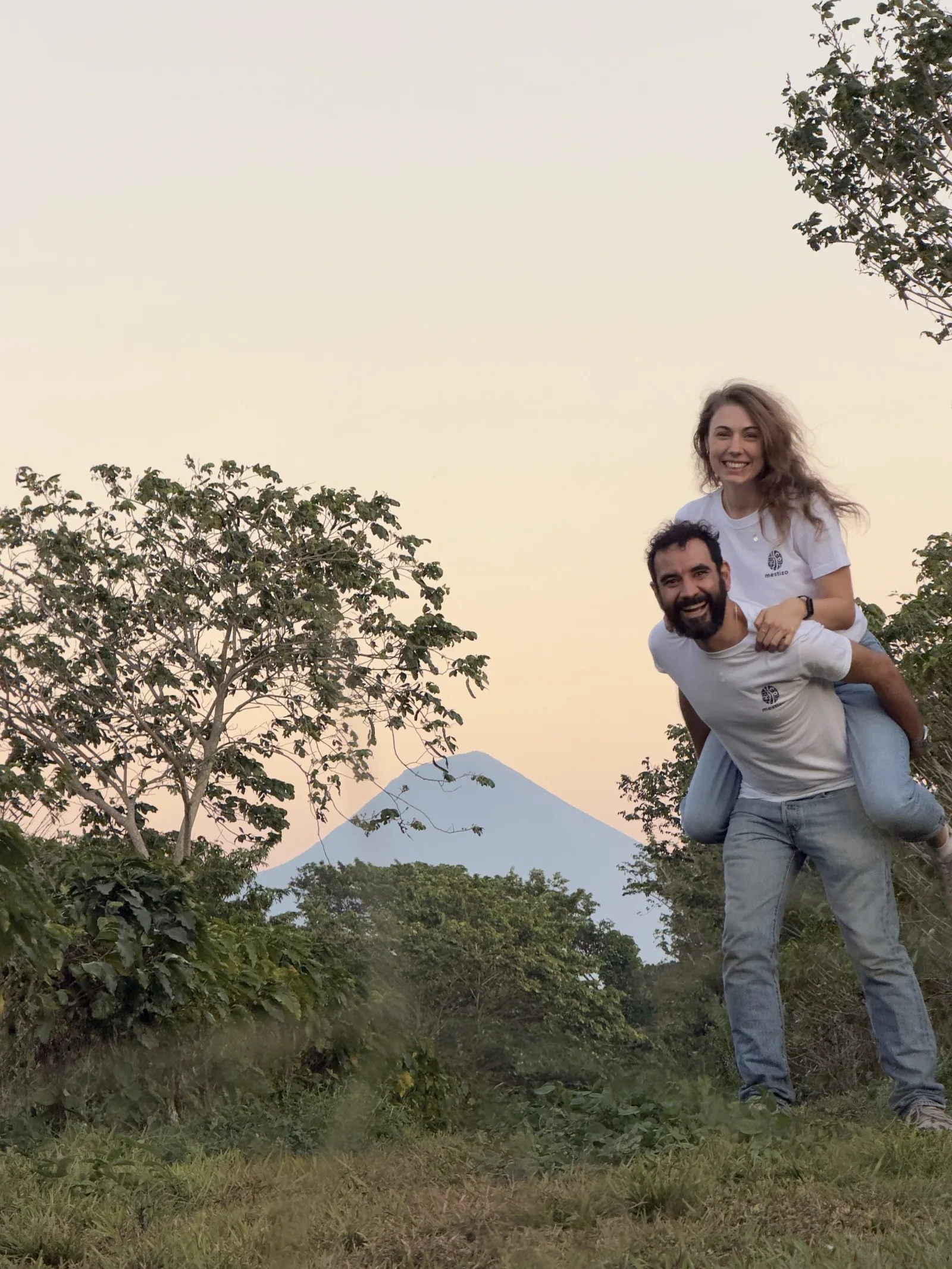 Joanna on Miguel's back, both laughing, a volcano in the distance behind them.